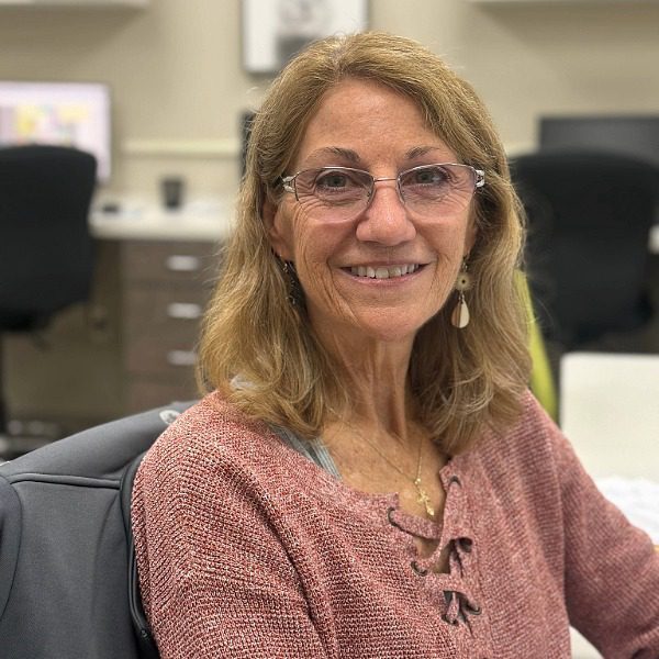Female with shoulder length dark blonde hair wearing glasses and a pink sir sitting at desk